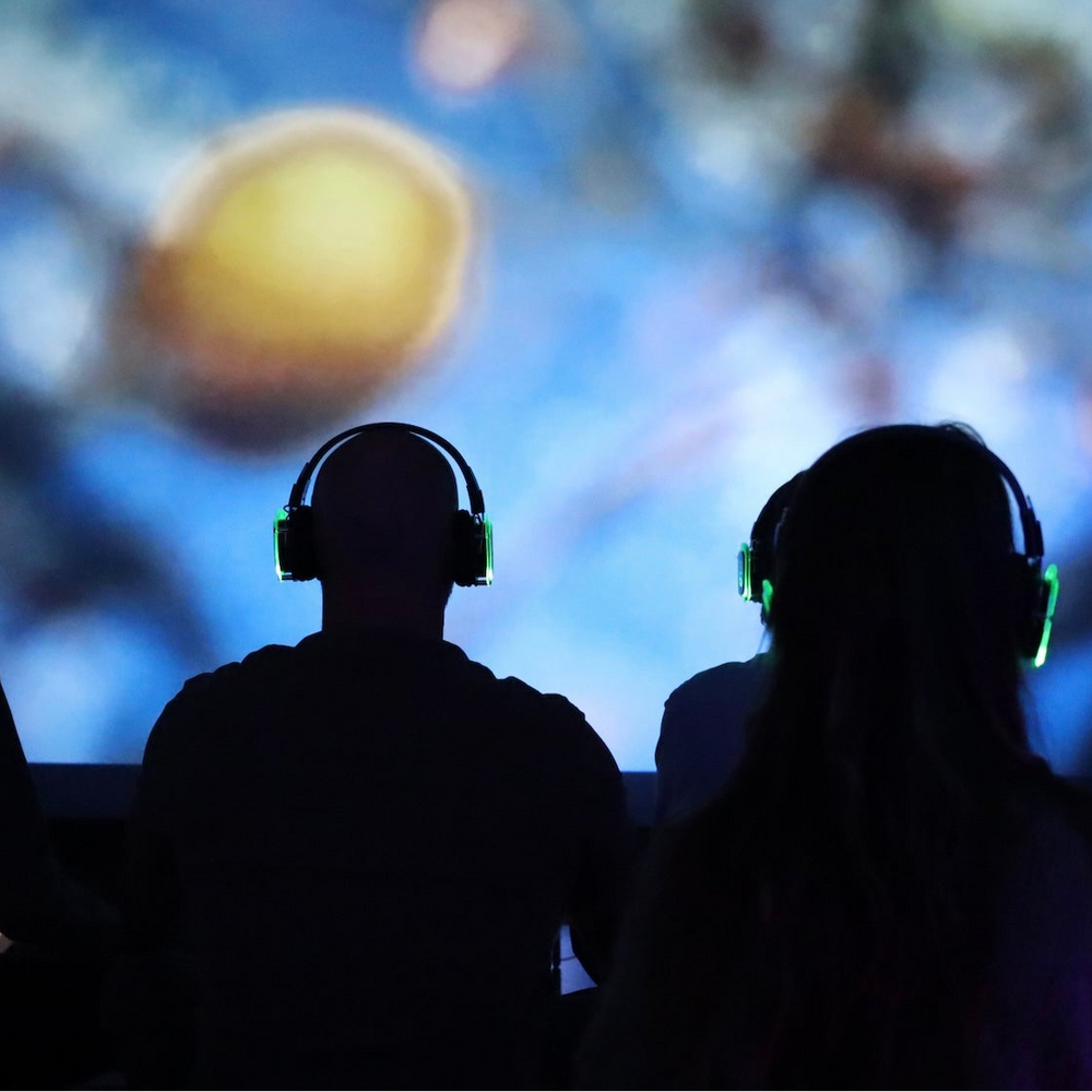 Silhouettes of people wearing headphones in front of a space-themed projection.
