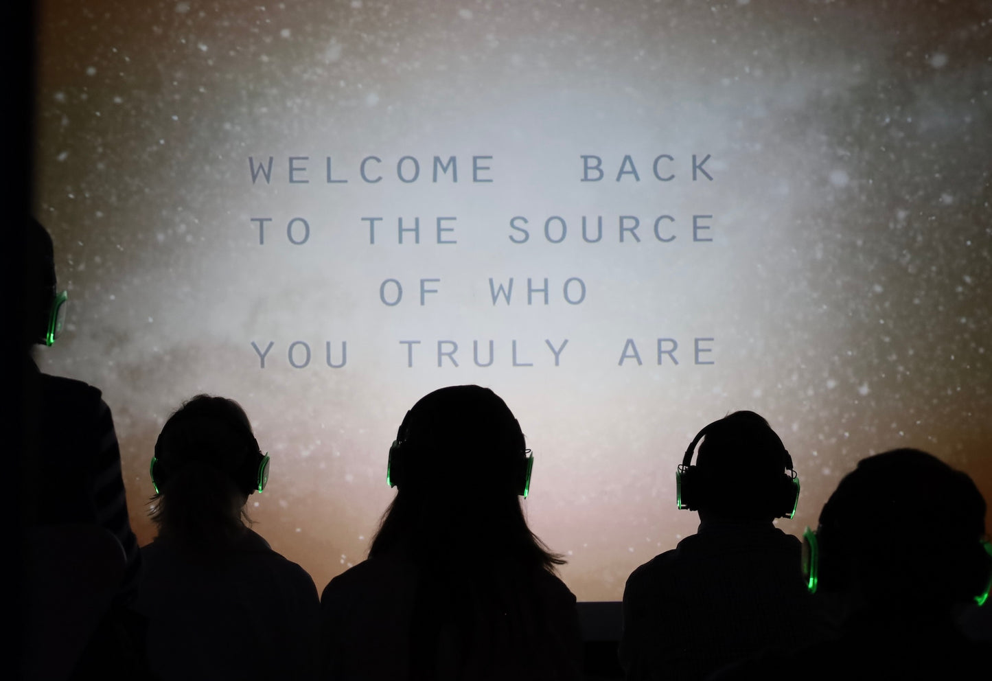 Silhouettes of people wearing headphones in front of a screen with text 'Welcome back to the source of who you truly are'.
